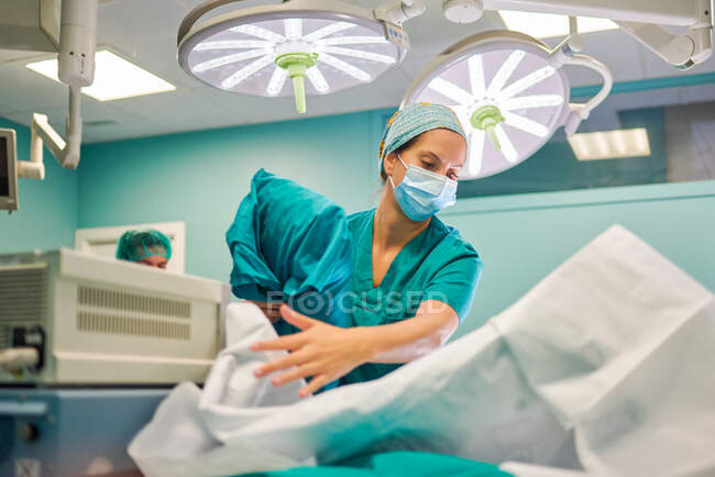 Dal basso assistente medico donna in uniforme preparare divano letto con lenzuola pulite in sala operatoria per la chirurgia in ospedale moderno — Foto stock