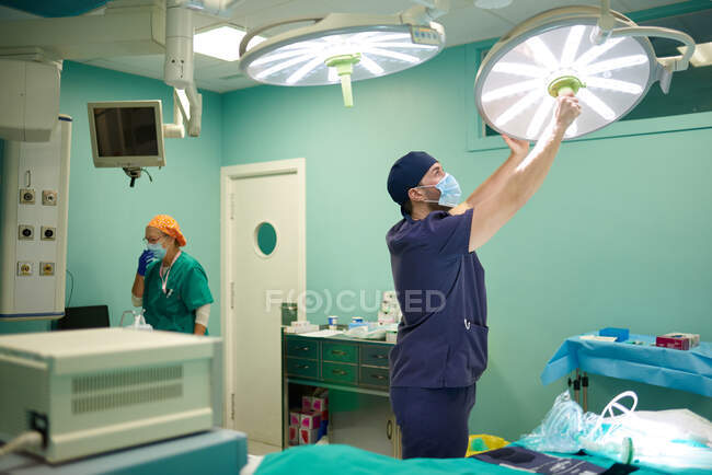 Концентрированный молодой медик в форме и маске, регулирующий хирургический свет перед операцией в современной больнице — стоковое фото