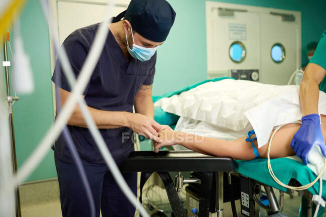 Anonymer junger Arzt in medizinischer Uniform und Maske legt intravenösen Katheter in die Hand eines gesichtslosen Patienten, der vor der Operation im Operationssaal auf der Couch liegt — Stockfoto