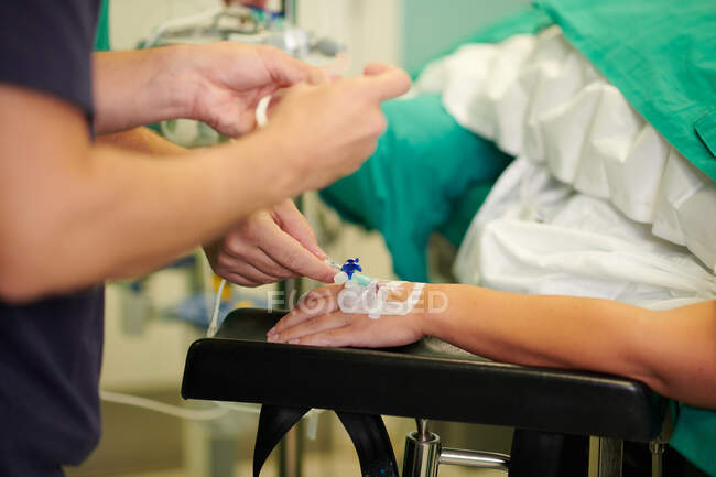 Crop unrecognizable medical assistants inserting intravenous catheter in hand on anonymous patient lying on couch in operating room — Stock Photo