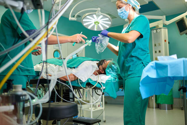 Vista laterale di assistenti femminili anonimi del raccolto in uniformi mediche e maschere che si preparano a dare l'anestesia al paziente sdraiato sul divano in sala operatoria — Foto stock