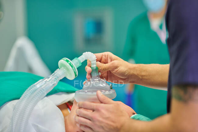 Anestesiologista masculino anônimo em uniforme médico e tampa segurando máscara de anestesia em face de paciente irreconhecível deitado no sofá na sala de cirurgia — Fotografia de Stock