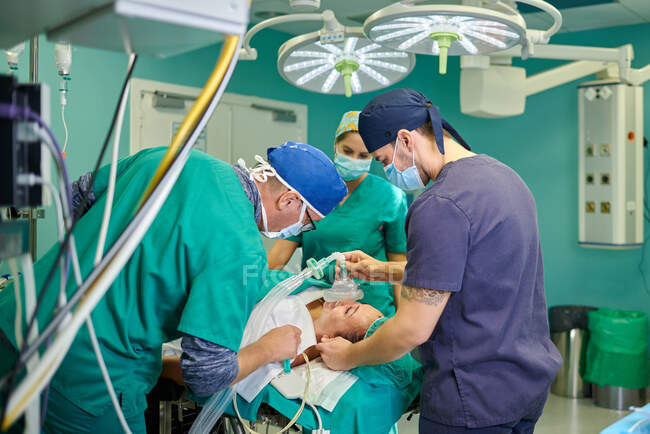 Vista lateral del anestesiólogo masculino anónimo en uniforme médico y gorra que sostiene la máscara de anestesia en la cara del paciente irreconocible acostado en el sofá en el quirófano sonriendo con el equipo de médicos - foto de stock