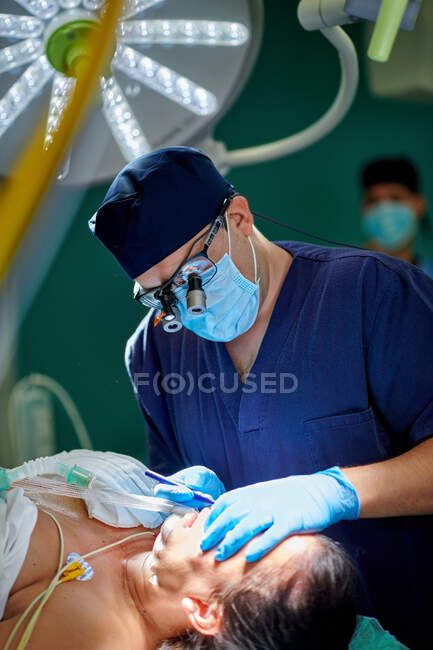 Unrecognizable male doctor in medical uniform and magnifying glasses performing rhinoplasty surgery for anonymous patient lying on couch in operating room — Stock Photo