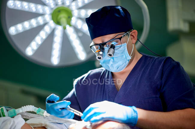 Médico masculino irreconhecível em uniforme médico e lupas realizando cirurgia de rinoplastia para paciente anônimo deitado no sofá na sala de cirurgia — Fotografia de Stock