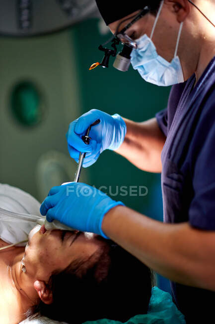 Visão lateral da cultura focada jovem cirurgião anônimo em uniforme médico e lentes de aumento fazendo injeção com seringa durante a realização de cirurgia de rinoplastia para paciente do sexo feminino — Fotografia de Stock