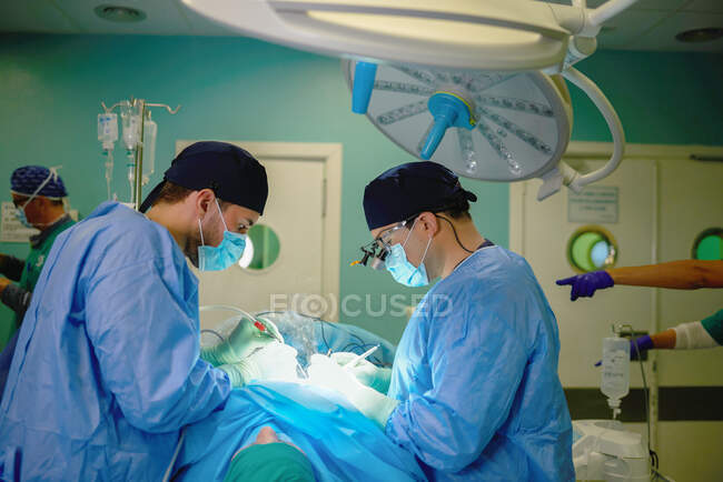 Vista lateral de un médico varón irreconocible con asistente en batas médicas y máscaras realizando cirugía con láser en quirófano - foto de stock