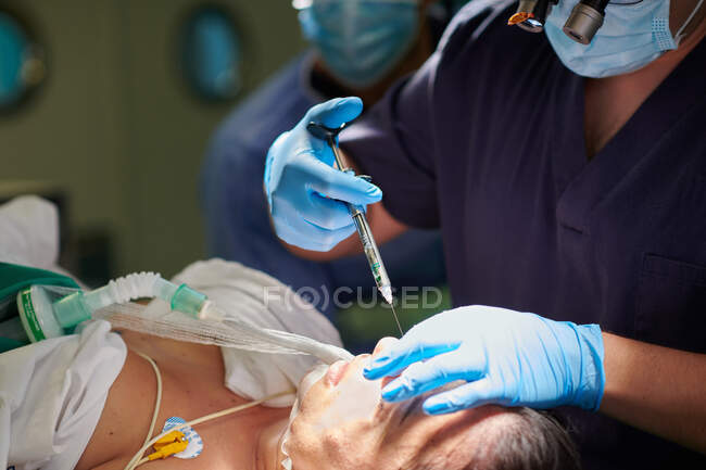 Crop focused anonymous young male surgeon in medical uniform and magnifying eyeglasses making injection with syringe while performing rhinoplasty operation for female patient — Stock Photo