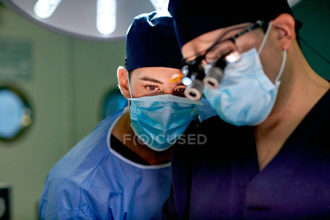 Jovem praticante masculino atencioso irreconhecível em vestido estéril e máscara após o processo de operação realizada pelo médico profissional na clínica moderna — Fotografia de Stock