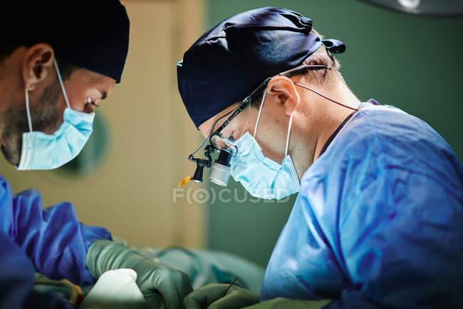 Vista lateral de un médico varón irreconocible con asistente en batas médicas y máscaras realizando cirugía con láser en quirófano - foto de stock