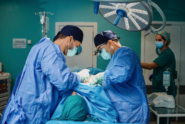 Paciente irreconhecível deitado na mesa de cirurgia durante a cirurgia executando por médicos do sexo masculino concentrados em vestidos médicos e máscaras — Fotografia de Stock