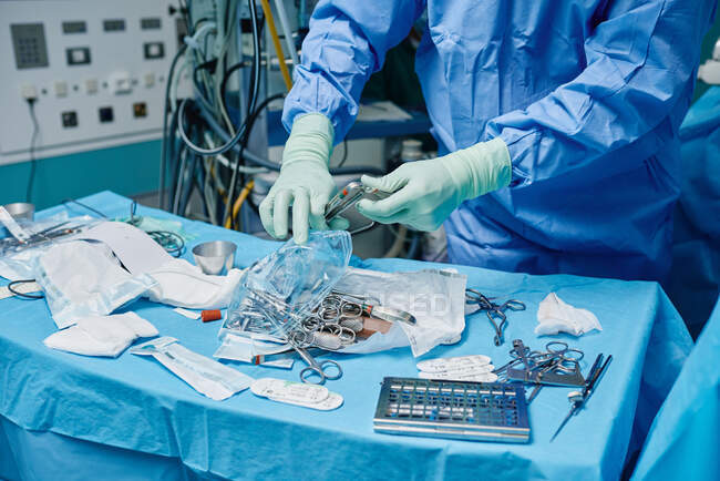 Crop anonymous surgeon in gown and latex gloves choosing various instruments scattered on sterile field while performing operation in hospital — Stock Photo
