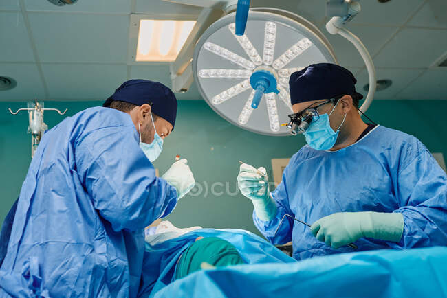 Paciente irreconocible acostado en la mesa de operaciones durante la cirugía realizado por médicos varones concentrados en batas y máscaras médicas - foto de stock