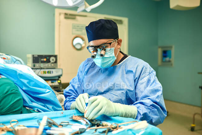Chirurgien masculin anonyme concentré en blouse bleue masque et lunettes grossissantes debout près d'un champ stérile avec divers outils médicaux pendant l'opération dans une clinique moderne — Photo de stock