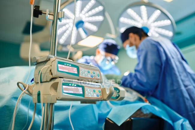 Von unten eine moderne Infusionspumpe im Operationssaal in der Nähe unkenntlich gemachter männlicher Ärzte, die in einem modernen Krankenhaus operieren — Stockfoto