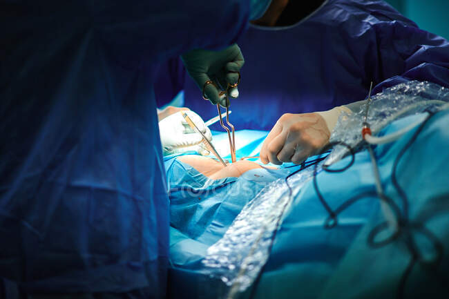 Cirujano anónimo de cultivo con asistente en batas estériles y guantes usando porta agujas mientras sutura la incisión durante la operación en la clínica moderna - foto de stock