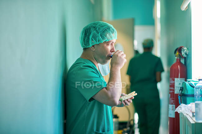 Side view of concentrated young male doctor in medical uniform and cap drinking takeaway coffee and eating sandwich after surgery — Stock Photo