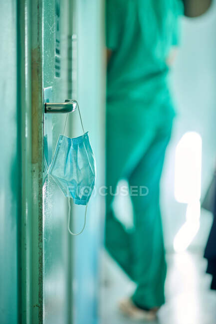Урожай неузнаваемый врач, стоящий в коридоре современной больницы рядом с дверью с медицинской маской висит на ручке — стоковое фото