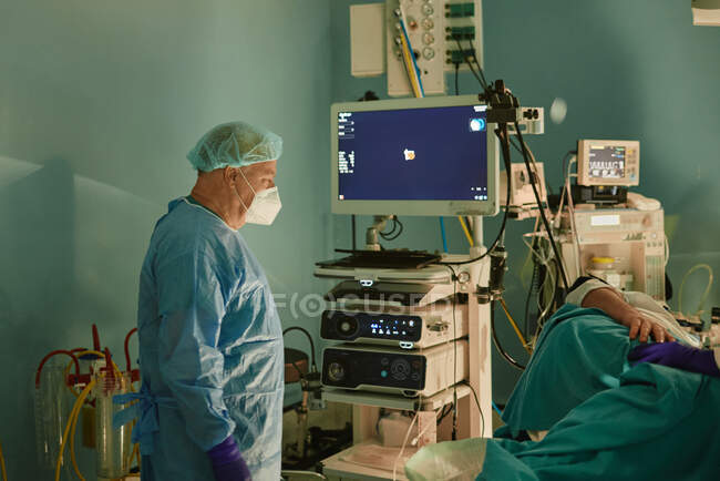 Побочный вид на неузнаваемого пожилого врача-мужчину в хирургическом халате и маске, стоящего рядом с пациентом, лежащим на диване перед эндоскопической процедурой — стоковое фото