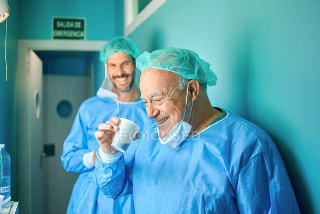 Cheerful senior and adult male surgeons in sterile gowns and caps  smiling and drinking takeaway coffee while standing in corridor after successful operation — Stock Photo
