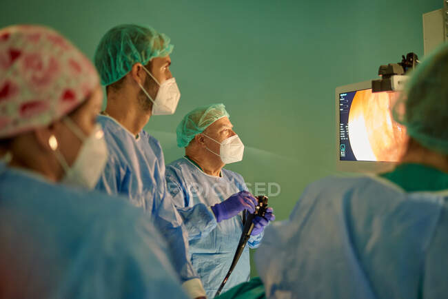 Група анонімних лікарів у хірургічних сукнях та масках, які дивляться на монітор під час вивчення пацієнта з ендоскопом перед операцією в сучасній клініці — стокове фото