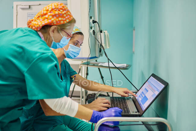 Side view of anonymous young females assistants in scrubs and masks using laptop during medical procedure in operating room — Stock Photo