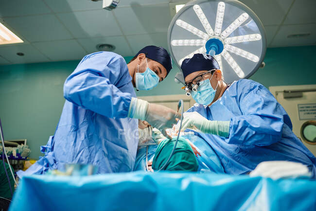 Снизу вид неузнаваемого молодого сконцентрированного ассистента-мужчины в стерильной маске и халате, дающего инструменты пластическому хирургу, выполняющему ринопластику в операционной — стоковое фото