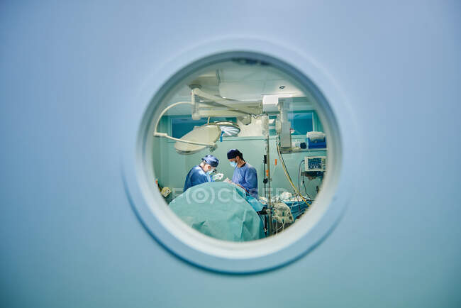 Durch das runde Fenster unkenntlich gemachter Ärzte in Uniformen und Masken, die im modernen Operationssaal operieren — Stockfoto