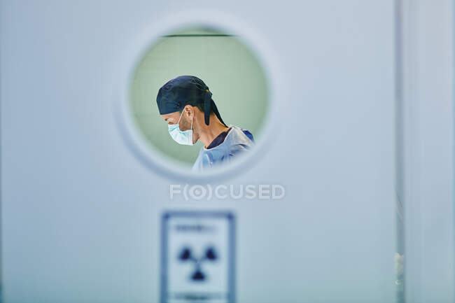 Побічний погляд на невідомого молодого зосередженого лікаря-чоловіка в медичній формі та маску, що стоїть в операційній кімнаті під час операції — стокове фото