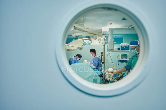 Через кругле вікно невизнаних лікарів у формі та масках, що виконують операцію в сучасній операційній кімнаті — стокове фото