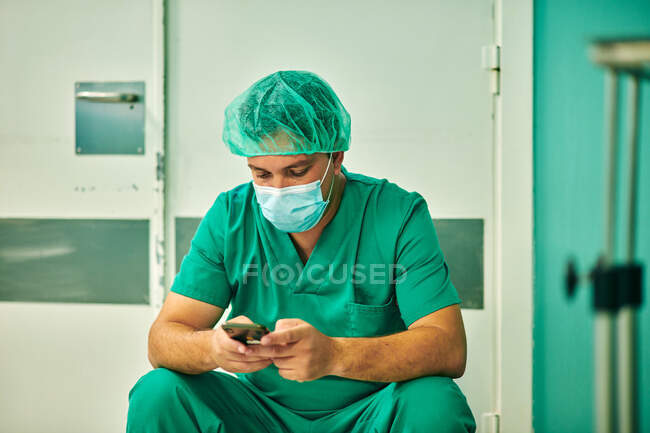 Anonymous young male doctor in green medical uniform and mask messaging on mobile phone while sitting in operating room after surgery — Foto stock