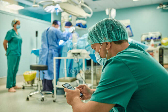 Seitenansicht eines anonymen, konzentrierten jungen männlichen Assistenten in medizinischer Uniform und Maskenbotschaft auf dem Handy, während ein Team professioneller Chirurgen in einer modernen Klinik operiert — Stockfoto