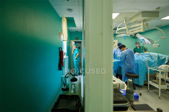 Вид сбоку анонимного практикующего мужчины в халате, разговаривающего по смартфону в коридоре, в то время как группа врачей проводит операцию в операционной — стоковое фото