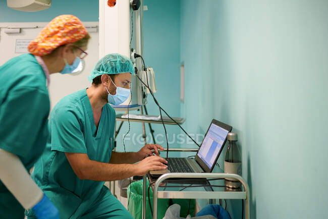 Side view of anonymous young female and male assistants in scrubs and masks using laptop during medical procedure in operating room — Foto stock