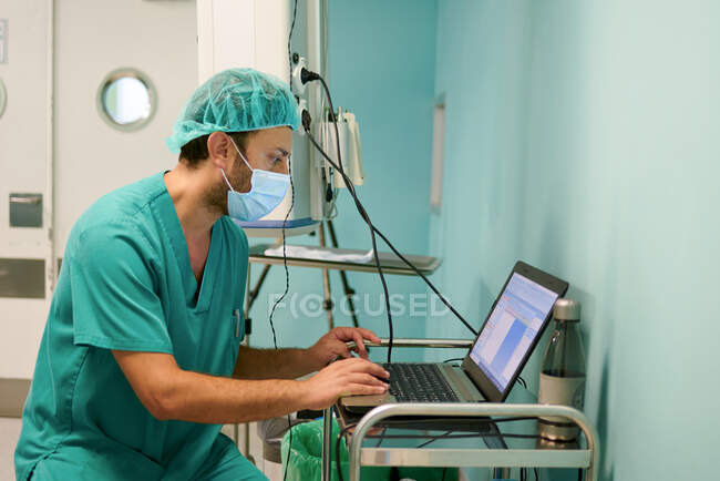 Seitenansicht des anonymen fokussierten jungen männlichen Assistenten in Peeling und Maske mittels Laptop während des medizinischen Eingriffs im Operationssaal — Stockfoto