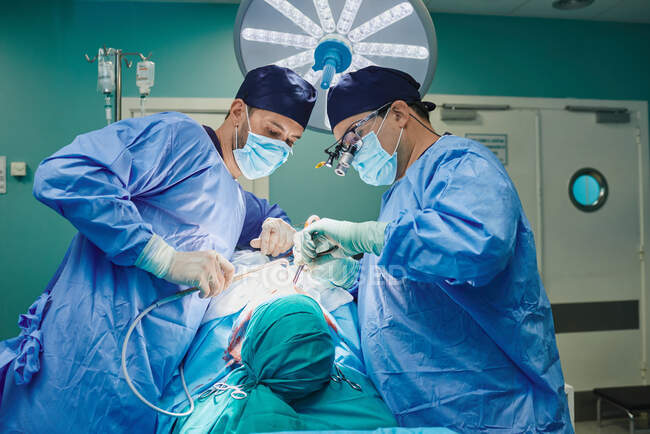 Seitenansicht eines unkenntlich gewordenen jungen konzentrierten männlichen Assistenten in steriler Maske und Mantel, der dem plastischen Chirurgen, der im Operationssaal Nasenkorrektur durchführt, Instrumente gibt — Stockfoto