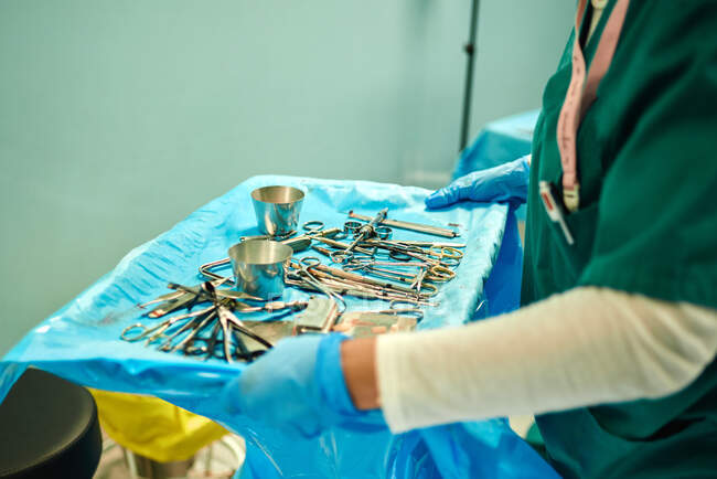 Crop anonymous assistant in latex gloves caring tray with various instruments scattered on sterile field while working operation in hospital — Fotografia de Stock