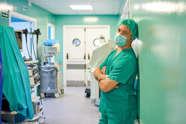 Side view of tired young male doctor in scrubs and medical mask leaning on wall with folded arms while resting in hospital corridor after surgery - foto de stock