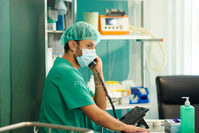 Vue latérale du jeune médecin masculin concentré en blouse et masque médical parlant au téléphone et regardant loin dans la salle d'hôpital — Photo de stock