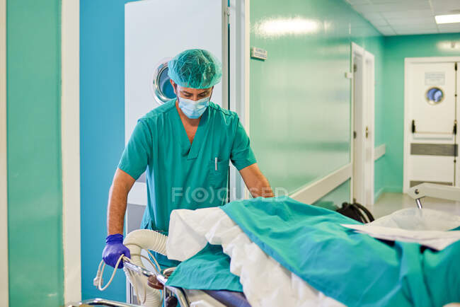 Неузнаваемый молодой практикующий мужчина в медицинской форме и маске, несущий анонимного пациента, лежащего на носилках в современном коридоре больницы — стоковое фото