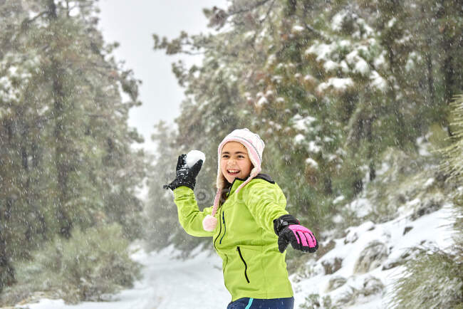 Захоплена дівчина в теплому одязі і капелюсі кидає сніжок, розважаючись у замерзлій зимовій лісі і дивлячись з щасливою посмішкою — стокове фото