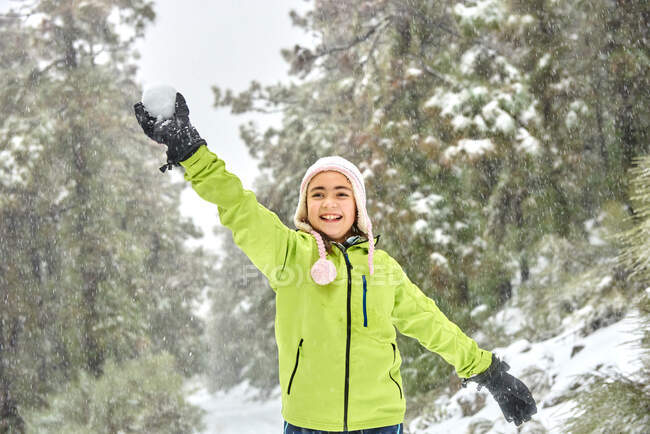 Excited girl in warm clothes and hat throwing snowball while having fun in frozen winter woodland and looking away with happy smile — Foto stock