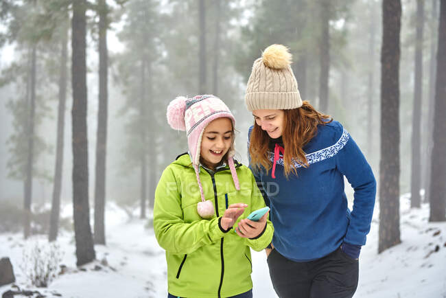Happy smiling mother and daughter in outerwear browsing mobile phone while walking together in snowy frozen woodland - foto de stock