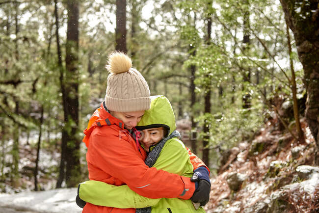 Side view happy smiling siblings in warm clothes embracing each other while spending peaceful winter day in snowy frozen woods - foto de stock