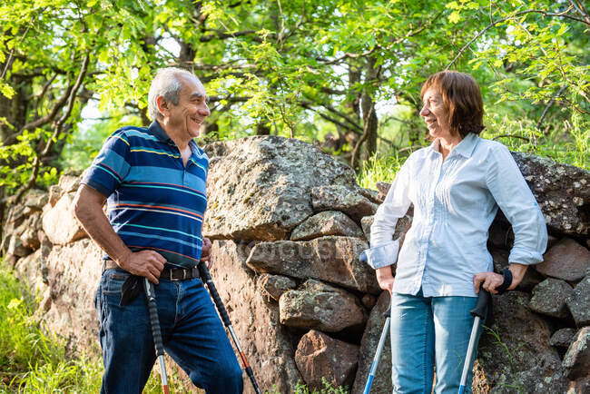 Cheerful mature couple standing on trail in green woods while having break during pole walking and looking at each other — Fotografia de Stock