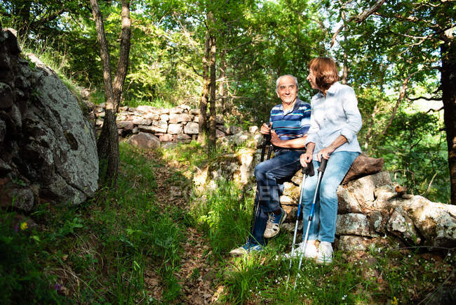 Cheerful mature couple standing on trail in green woods while having break during pole sitting and looking at each other — Stock Photo