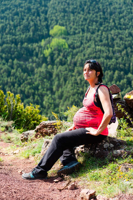 Pregnant female traveler sitting on mossy stones in green woods and having break during trekking in summer — Stock Photo