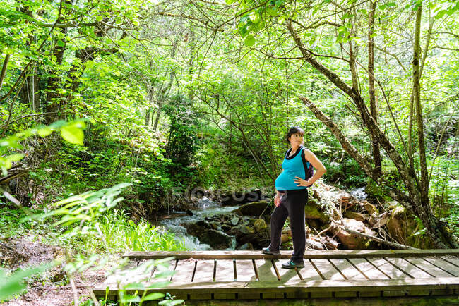 Vue latérale de touriste femelle enceinte debout sur un pont en bois dans les bois et profiter de la nature pendant les vacances d'été sur fond de rivière rapide — Photo de stock