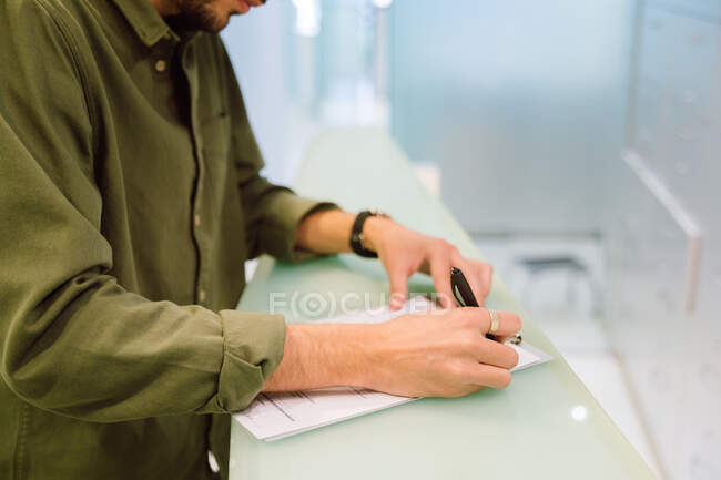 Side view crop focused male in casual clothes filling survey form while standing near clinic reception counter — Fotografia de Stock