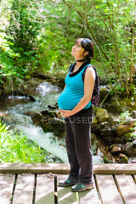 Seitenansicht einer lächelnden schwangeren Touristin, die auf einer Holzbrücke im Wald steht und die Natur im Sommerurlaub vor dem Hintergrund des schnellen Flusses genießt — Stockfoto